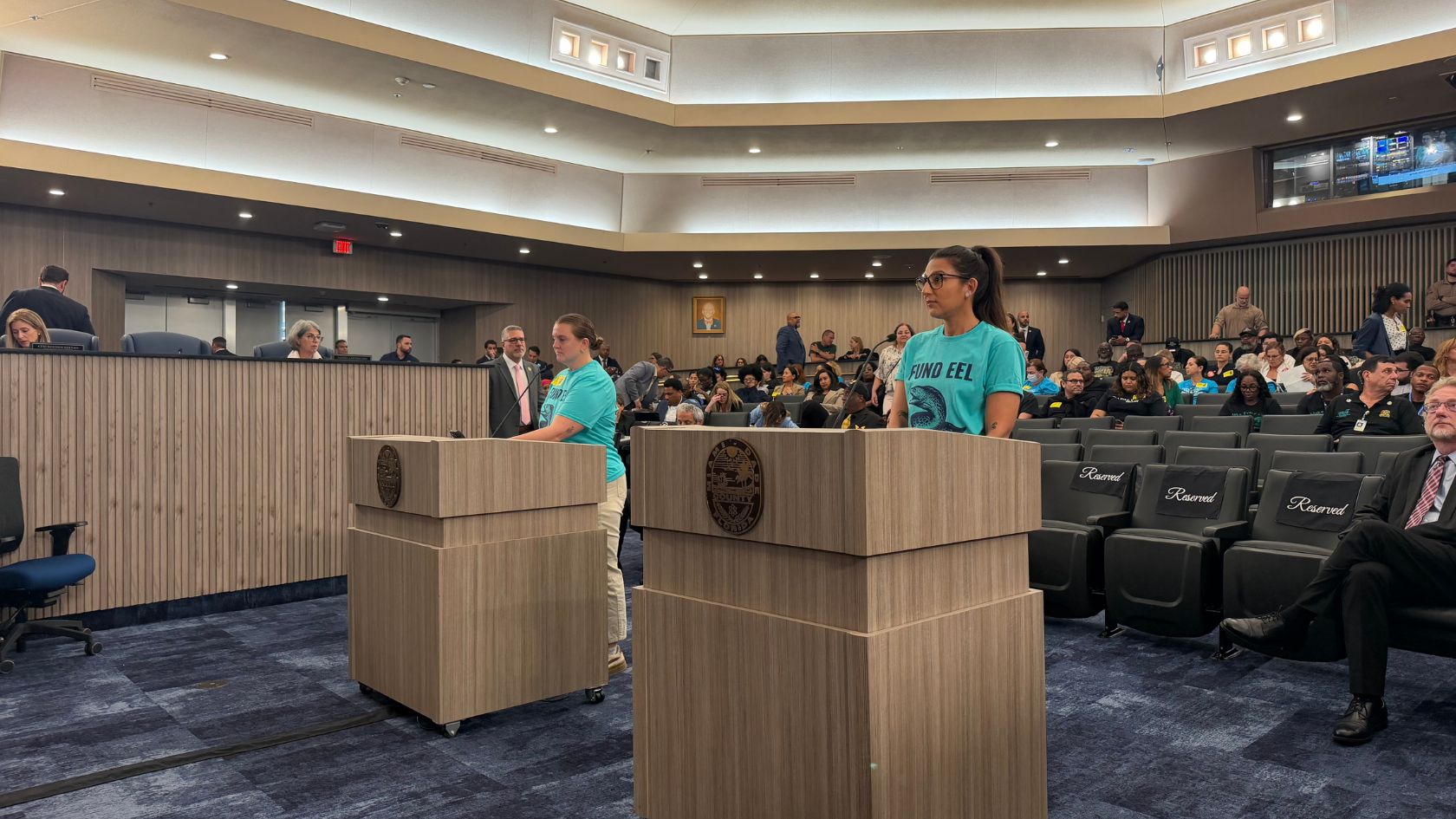 Advocates in teal FUND EEL shirts give public comment at podiums in a packed county commission chamber.