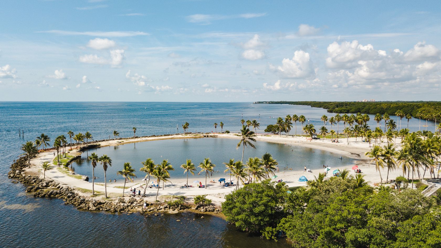 Aerial view of a circular atoll swimming lagoon ringed by palms and a white sand beach on a shallow bay.