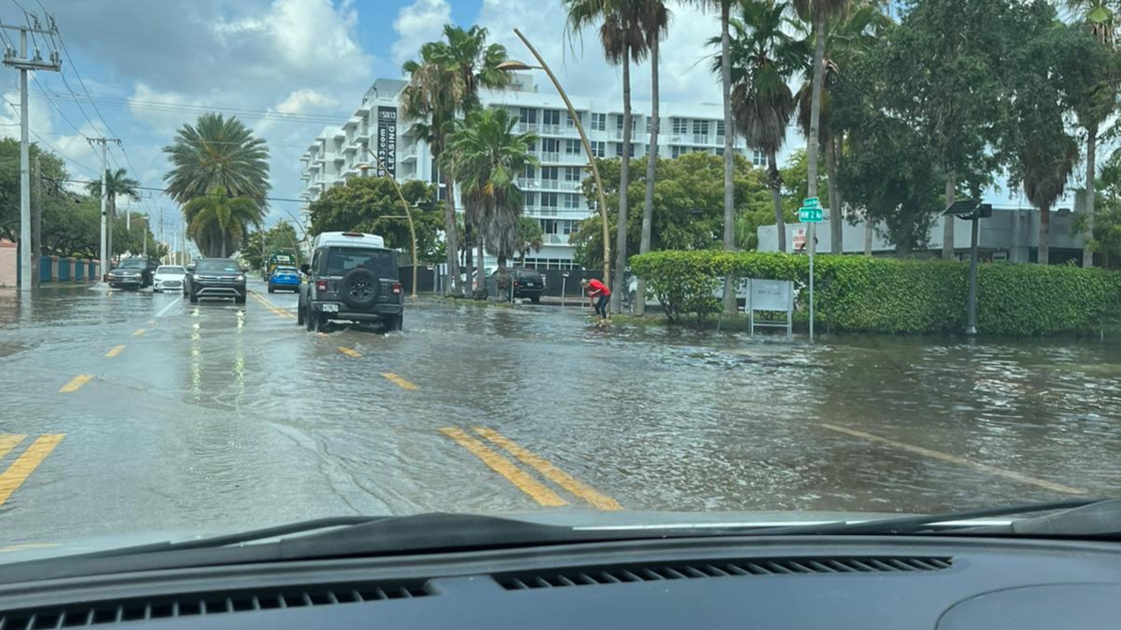Vehicles drive through a flooded street while a worker in a red shirt clears a storm drain at the curb.