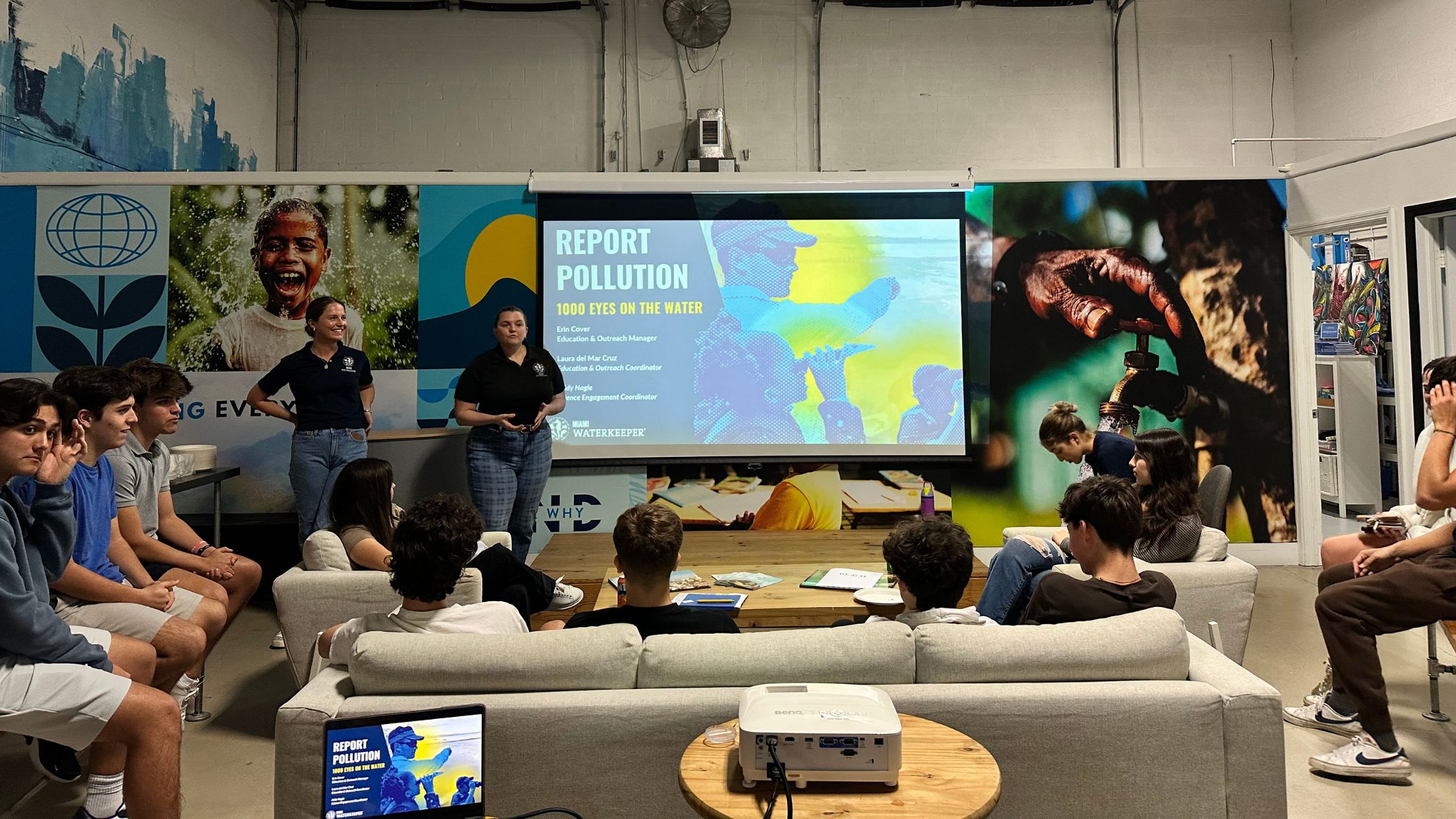 Two Miami Waterkeeper educators present the Report Pollution: 1000 Eyes on the Water slide to students seated on couches.