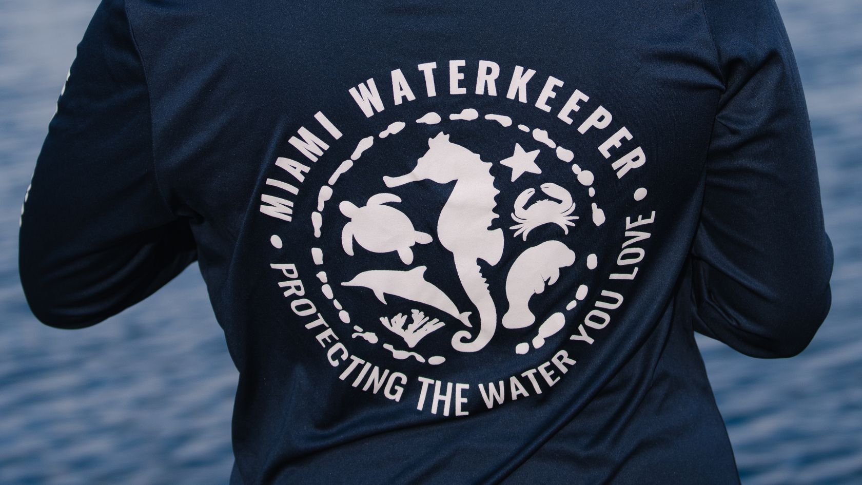 Close-up of a navy Miami Waterkeeper shirt showing the seahorse seal and the tagline Protecting the Water You Love.