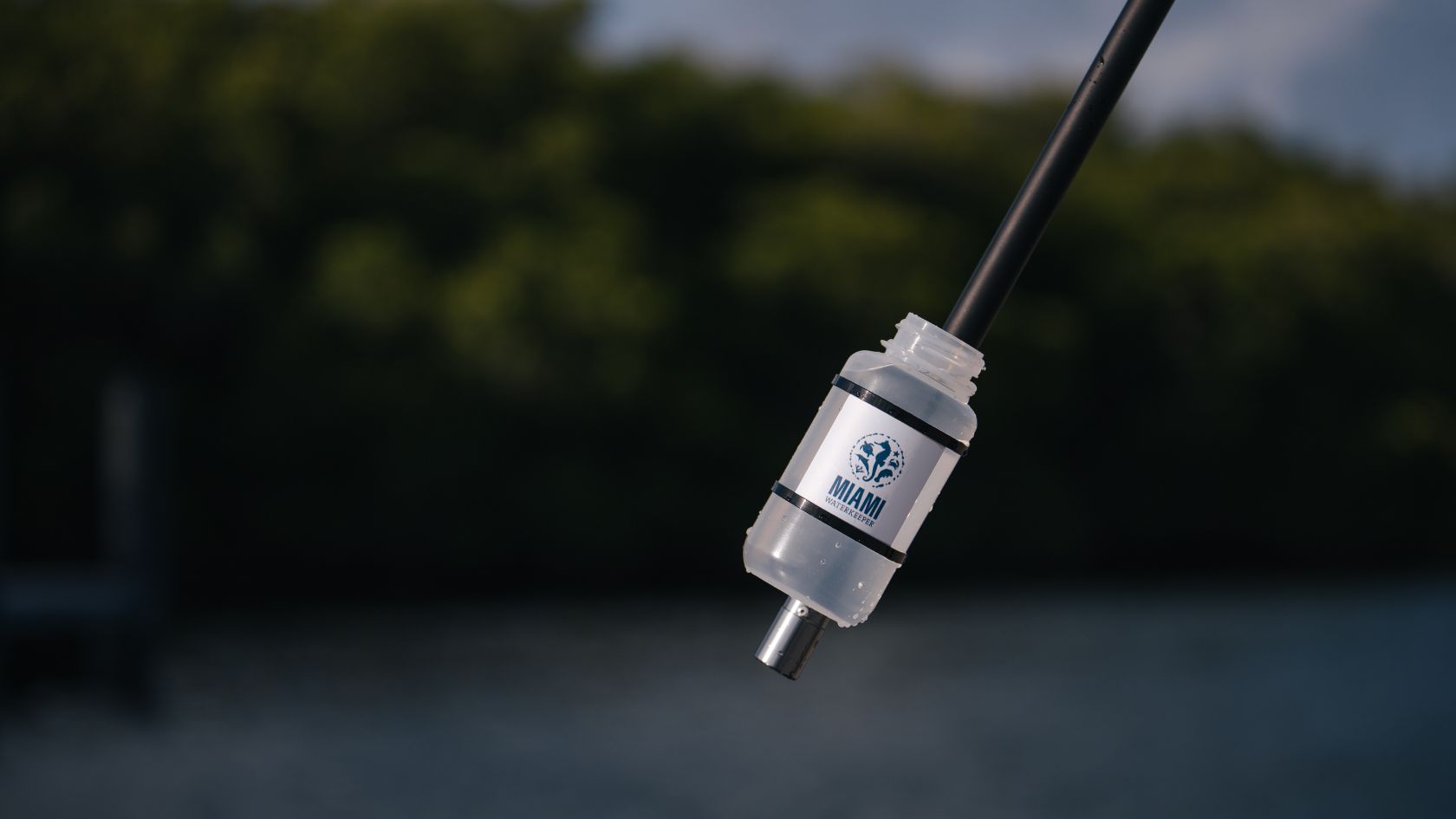 A Miami Waterkeeper labeled sample bottle mounted on a sampling pole above the water, mangroves behind.
