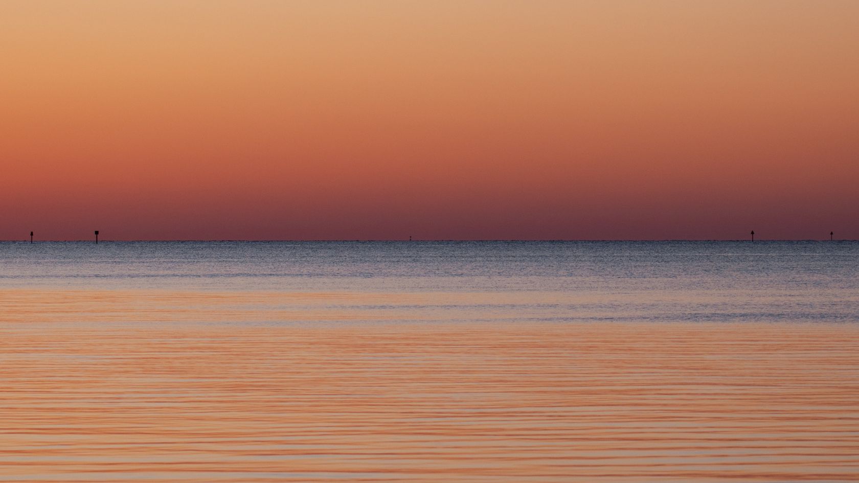 Orange and pink sunset light over calm rippled water with distant channel markers on the horizon.